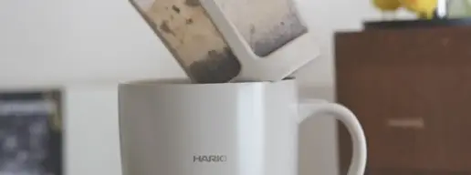 Hands pour loose tea leaves from a dual chamber infuser into a white HARIO mug The scene sits on a checkered tablecloth with a wooden cabinet in the background