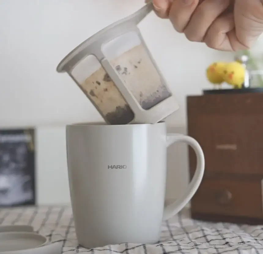 Hands pour loose tea leaves from a dual chamber infuser into a white HARIO mug The scene sits on a checkered tablecloth with a wooden cabinet in the background
