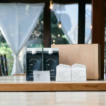 Two dark blue cartons with white caps on a wooden table surrounded by a brown box and printed info cards in a sunlit indoor space with sheer curtains