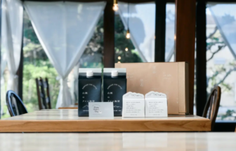 Two dark blue cartons with white caps on a wooden table, surrounded by a brown box and printed info cards in a sunlit indoor space with sheer curtains.