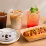 Spread of beverages and a hot dog on a wooden table iced coffee iced latte pink grapefruit drink a hot dog with toppings and a bear shaped cookie on a plate