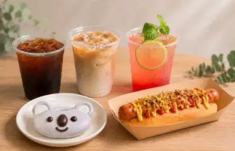 Spread of beverages and a hot dog on a wooden table iced coffee iced latte pink grapefruit drink a hot dog with toppings and a bear shaped cookie on a plate