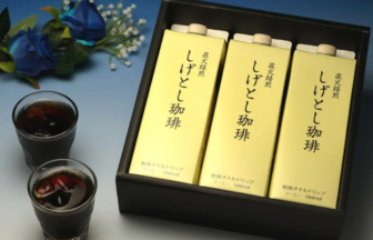 Three yellow coffee cartons in a black gift box, with two glasses of iced coffee and blue flowers nearby.