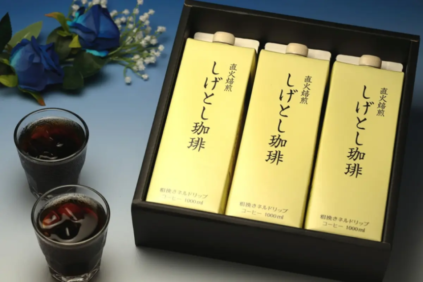 Three yellow coffee cartons in a black gift box with two glasses of iced coffee and blue flowers nearby