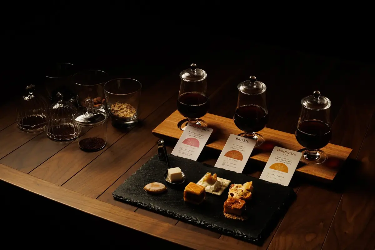 暗い木目調のテーブルに、3種類のコーヒーテイスティングセットが並べられています。それぞれのコーヒーには産地と特徴が書かれたカードが添えられ、スレートプレートにはコーヒーペアリング用のお菓子が数種類置かれています。高級感のあるコーヒー体験を演出しています。
