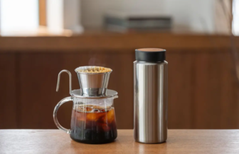 Pour over coffee setup on a wooden counter glass carafe with a metal dripper and filter next to a stainless steel thermos