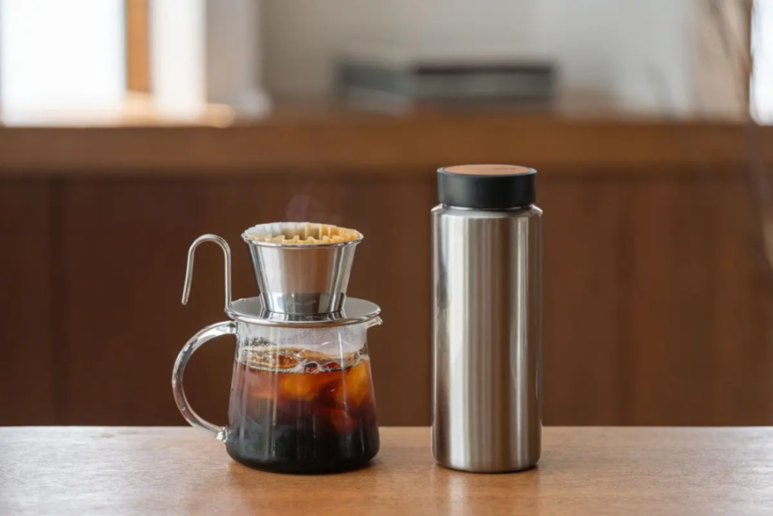 Pour over coffee setup on a wooden counter glass carafe with a metal dripper and filter next to a stainless steel thermos
