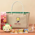 Beige tote bag with a Coji Coji logo on a wooden surface surrounded by snacks a plush lion toy a honey peach juice carton and a glass