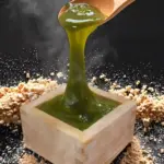 Green thick sauce being poured into a wooden square cup with crumbled bits scattered around on a dark surface