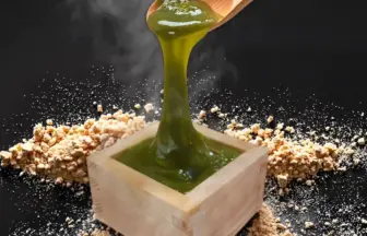 Green thick sauce being poured into a wooden square cup with crumbled bits scattered around on a dark surface