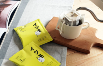 Pour over coffee setup with a white mug and filter resting on a wooden board and two bright yellow coffee packets nearby on a light table