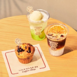 Green lime drink in a plastic cup iced coffee with an orange slice and a blueberry muffin on a wooden table at The Coach Coffee Shop napkin shows logo