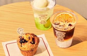 Green lime drink in a plastic cup, iced coffee with an orange slice, and a blueberry muffin on a wooden table at The Coach Coffee Shop (napkin shows logo).