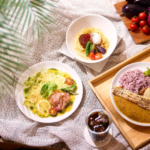 Overhead view of a meal spread lemon chicken pasta a curry rice plate with a breadstick and a bowl of pasta with tomatoes and greens on a wooden tray with iced coffee nearby