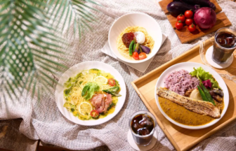 Overhead view of a meal spread lemon chicken pasta a curry rice plate with a breadstick and a bowl of pasta with tomatoes and greens on a wooden tray with iced coffee nearby