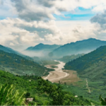 Winding river through a lush green valley flanked by misty mountains under a cloudy blue sky
