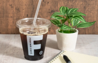 Iced coffee in a clear plastic cup with a straw on a white desk, beside a small potted plant and a spiral notebook with a pen.
