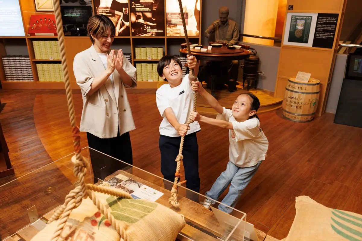 博物館で、女性と2人の子供がコーヒーの展示を楽しんでいる。子供たちはロープを引く体験型展示に笑顔で挑戦し、女性は拍手で見守る。ジャマイカ産ブルーマウンテンコーヒーの樽も見える。