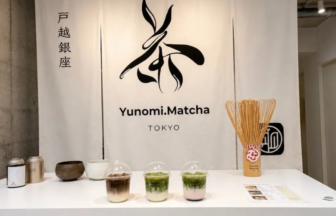 Tea counter with matcha drinks in clear cups a bamboo whisk and a white backdrop reading YunomiMatcha Tokyo