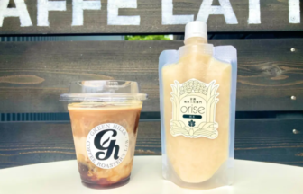 Iced coffee in a clear cup with lid beside a beige refill pouch labeled orise on a white table against a wooden fence backdrop