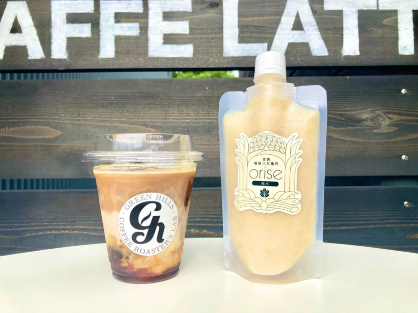 Iced coffee in a clear cup with lid beside a beige refill pouch labeled orise on a white table against a wooden fence backdrop