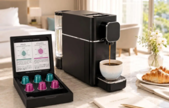 Compact black espresso machine brews into a white mug on a marble countertop with a croissant and newspaper nearby in a bright hotel room