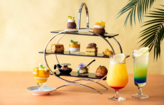 Three tier dessert stand with assorted pastries and small desserts three colorful drinks on a peach table with a tropical plant in the background