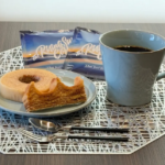 A blue mug of black coffee beside a plate with a ring shaped pastry and a bite taken out on a decorative placemat with two snack packages in the background