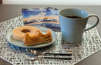 A blue mug of black coffee beside a plate with a ring shaped pastry and a bite taken out on a decorative placemat with two snack packages in the background