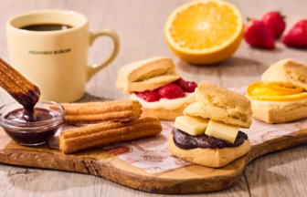 Assorted breakfast pastries on a wooden board churros with chocolate dip jam pastries a cheese sandwich orange half strawberries and a mug of coffee