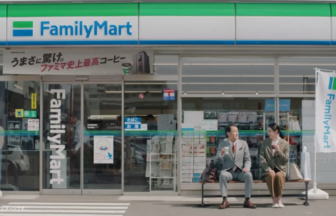Two people in business attire sit on a bench outside a FamilyMart storefront, chatting over drinks.