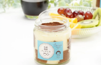 Layered dessert in a glass jar with cocoa powder top, blue label, and fruit platter in the background.