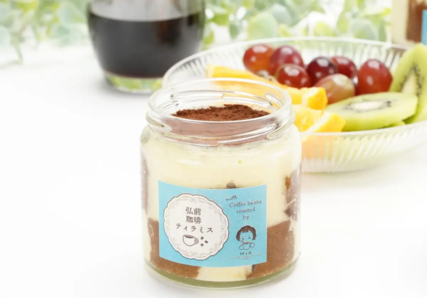 Layered dessert in a glass jar with cocoa powder top blue label and fruit platter in the background