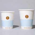 Two white disposable coffee cups with a light blue band and a brown octagonal brand badge placed on a neutral surface
