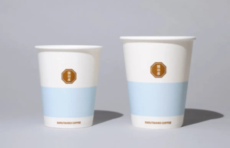 Two white disposable coffee cups with a light blue band and a brown octagonal brand badge placed on a neutral surface