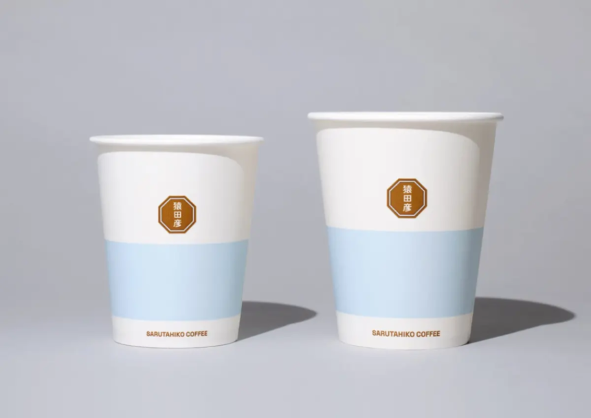Two white disposable coffee cups with a light blue band and a brown octagonal brand badge placed on a neutral surface