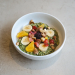 White bowl with a green smoothie bowl topped with banana slices orange segments berries and chopped nuts on a gray surface close up