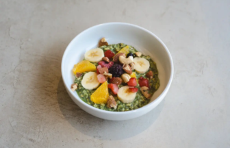 White bowl with a green smoothie bowl topped with banana slices, orange segments, berries, and chopped nuts on a gray surface, close-up.