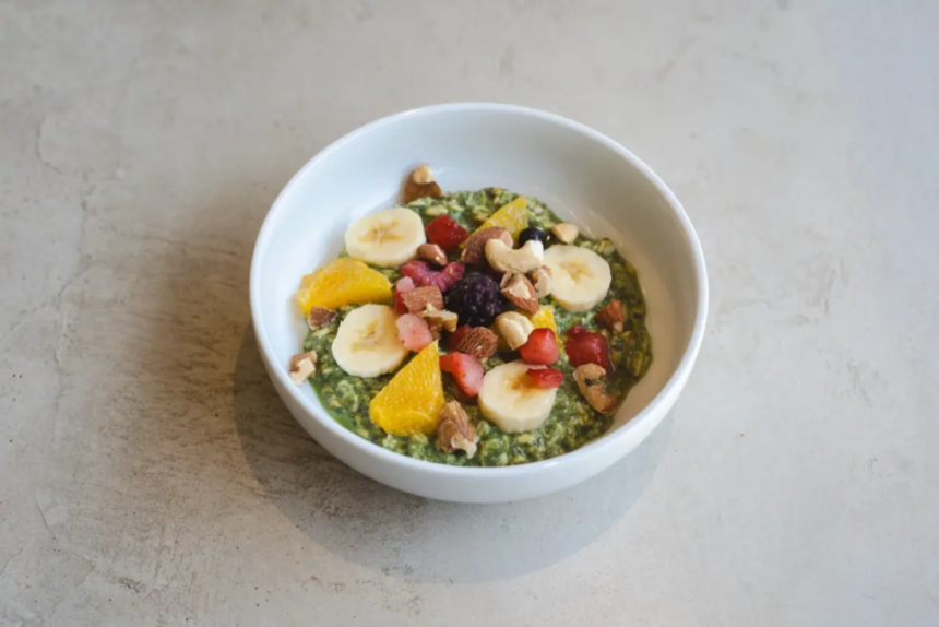 White bowl with a green smoothie bowl topped with banana slices orange segments berries and chopped nuts on a gray surface close up