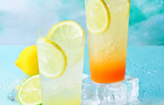 Two tall citrus drinks with lemon wedges and ice set on a blue icy surface with lime whole lemon and mango cubes nearby