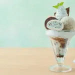 Ice cream sundae in a tall glass with mint leaf cookies and chocolate pieces on a pale green background