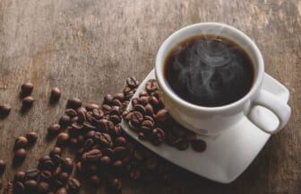Cup of hot black coffee with steam on a white saucer, surrounded by roasted beans on a rustic wooden table, close-up view.