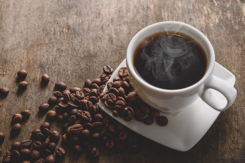Cup of hot black coffee with steam on a white saucer surrounded by roasted beans on a rustic wooden table close up view