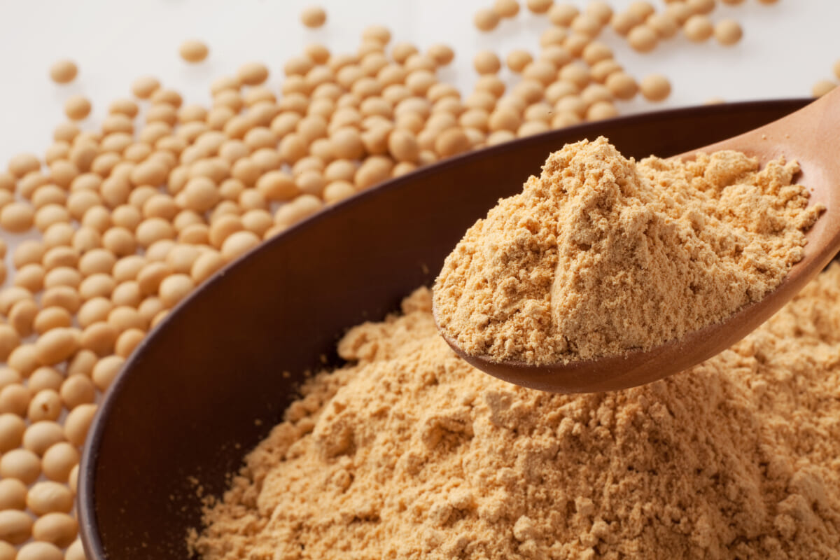 Close up of a wooden spoon lifting light brown peanut powder from a bowl with whole peanuts scattered in the background