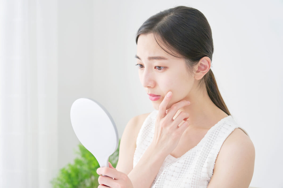 Woman in a white top looks at her reflection in a handheld mirror touching her cheek by a bright window