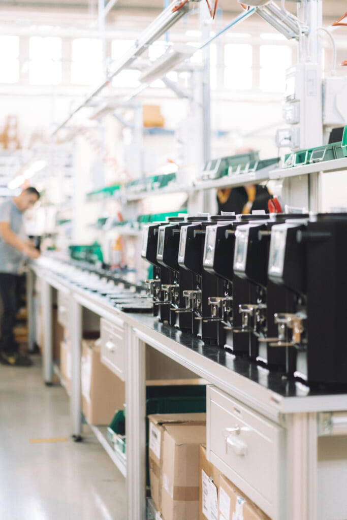 Row of black electronic devices on a factory workbench with boxes stored beneath a blurred technician in the background on the left