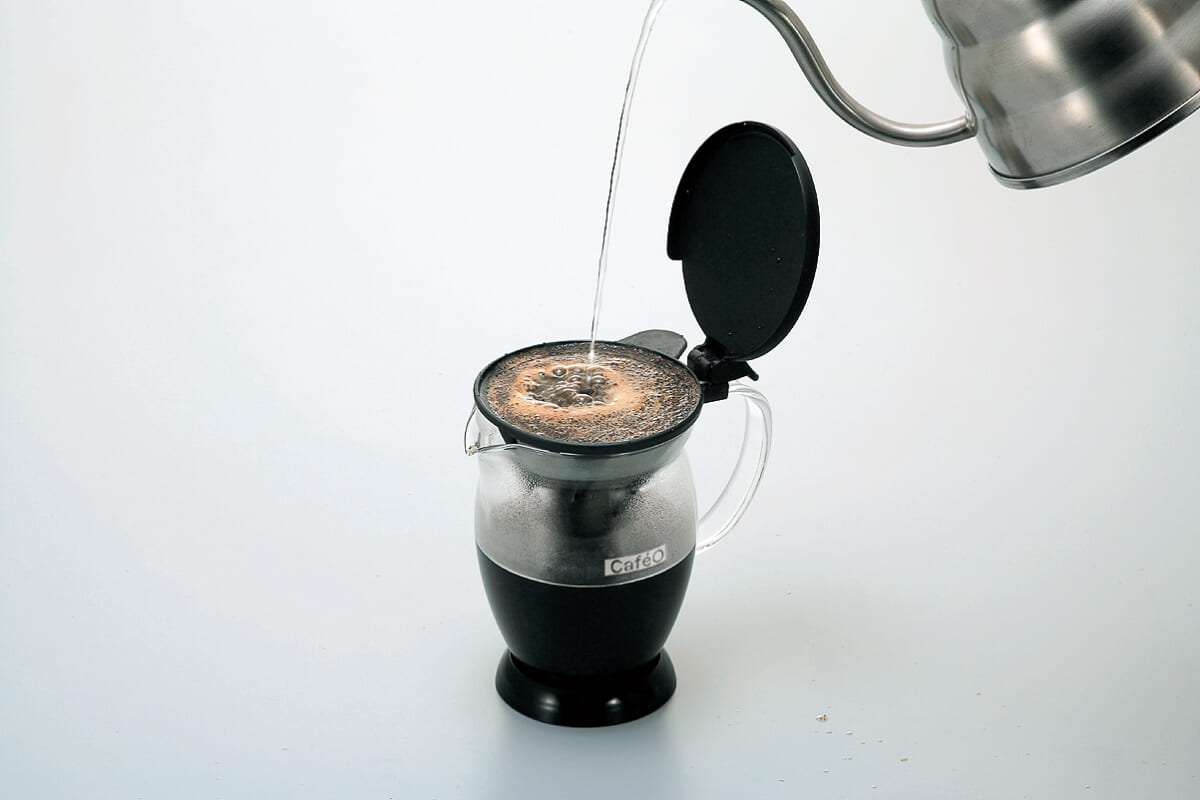 A moka pot on a white surface with hot water being poured into its top chamber from a kettle before brewing coffee