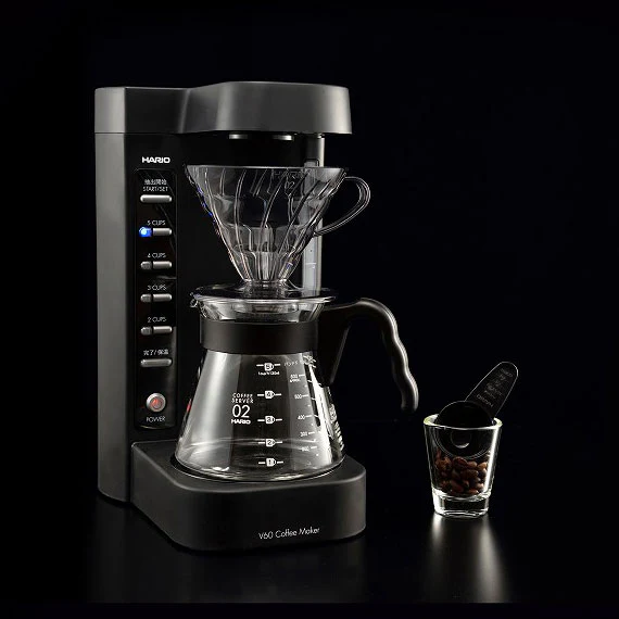 Hario V60 drip coffee maker with glass carafe and cone set on a dark surface next to a small glass with coffee beans nearby