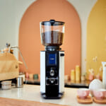 Commercial coffee grinder with glass hopper filled with beans on a white countertop in a bright cafe setting