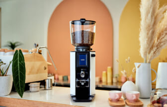 Commercial coffee grinder with glass hopper filled with beans on a white countertop in a bright cafe setting